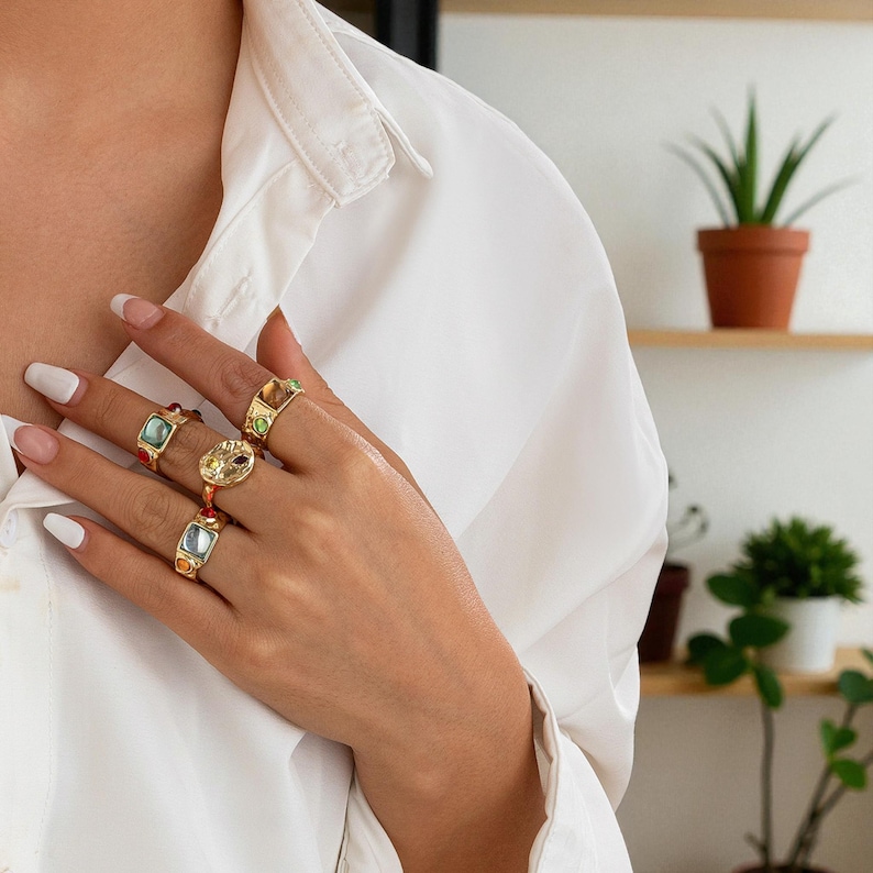 May include: A hand wearing several gold rings with colorful gemstones. The rings feature various shapes and colors, including red, blue, green, and yellow. The rings are set against a white shirt.