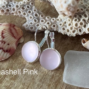 May include: A pair of silver hoop earrings with a light pink glass cabochon. The earrings are displayed on a wooden surface with seashells and sea glass.