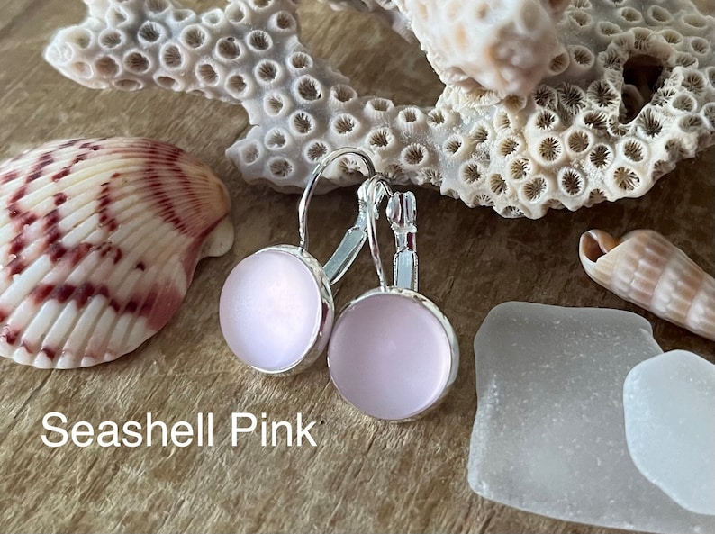 May include: A pair of silver hoop earrings with a light pink glass cabochon. The earrings are displayed on a wooden surface with seashells and sea glass.