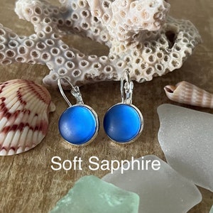 May include: A pair of silver dangle earrings with a soft blue round glass cabochon. The earrings are displayed on a wooden surface with seashells and sea glass. The text "Soft Sapphire" is visible below the earrings.