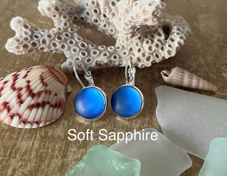 May include: A pair of silver dangle earrings with a soft blue round glass cabochon. The earrings are displayed on a wooden surface with seashells and sea glass. The text "Soft Sapphire" is visible below the earrings.