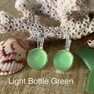 May include: A pair of light bottle green glass earrings with silver hooks. The earrings are round and have a smooth, frosted finish. The earrings are displayed on a wooden surface with seashells and sea glass.