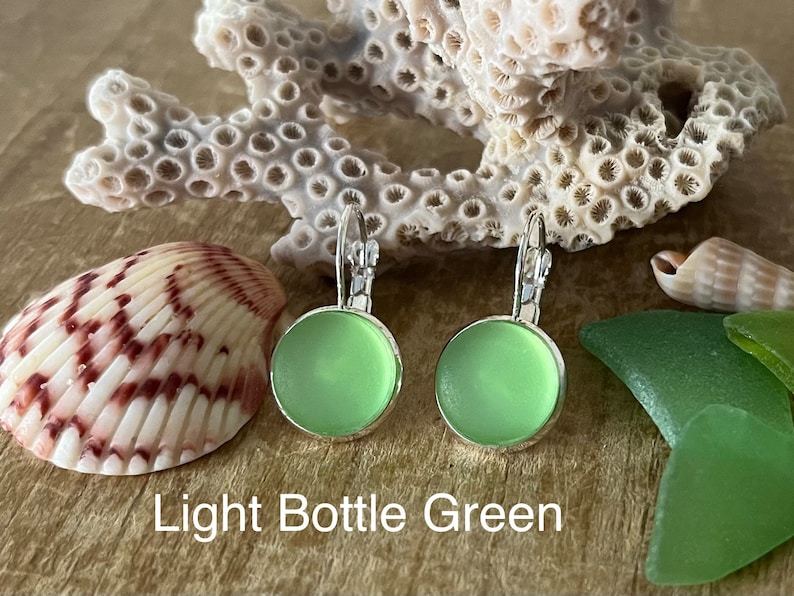 May include: A pair of light bottle green glass earrings with silver hooks. The earrings are round and have a smooth, frosted finish. The earrings are displayed on a wooden surface with seashells and sea glass.