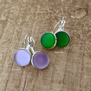 May include: Two pairs of earrings with silver hooks and round glass stones. One pair has lavender stones and the other pair has green stones. The text "Lavender" and "Shamrock" are written below the earrings.