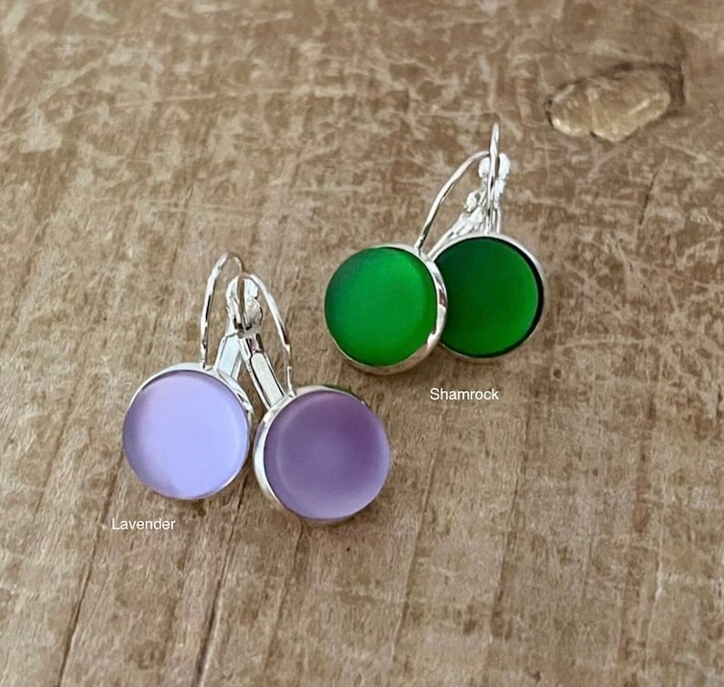 May include: Two pairs of earrings with silver hooks and round glass stones. One pair has lavender stones and the other pair has green stones. The text "Lavender" and "Shamrock" are written below the earrings.