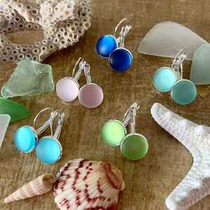 May include: Six pairs of silver dangle earrings with round glass stones in various pastel colours, including blue, green, pink, and light blue. The earrings are displayed on a wooden surface with seashells and sea glass.