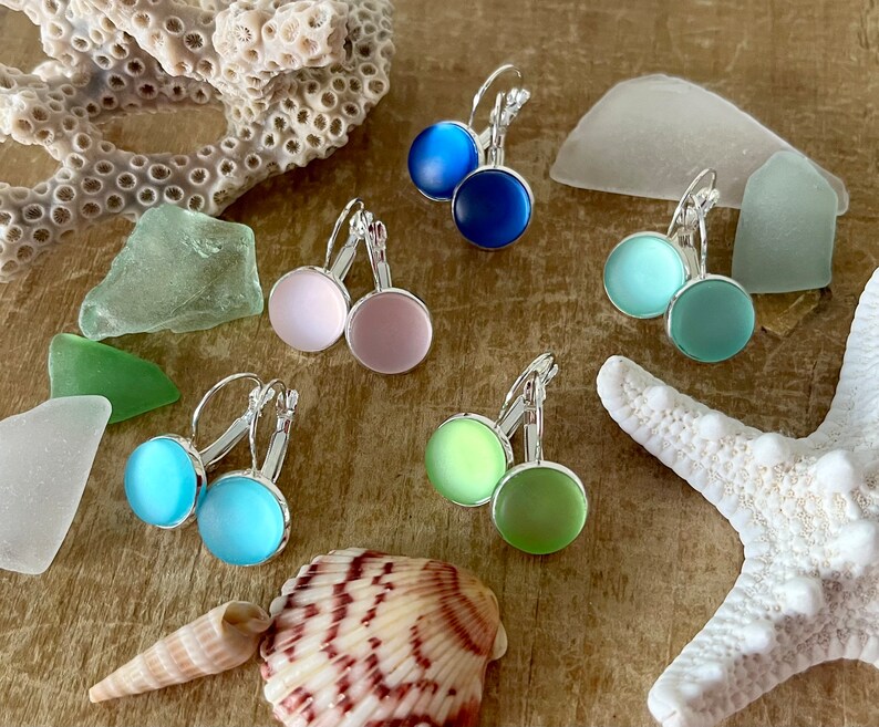 May include: Six pairs of silver dangle earrings with round glass stones in various pastel colours, including blue, green, pink, and light blue. The earrings are displayed on a wooden surface with seashells and sea glass.