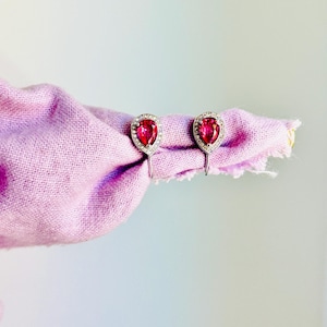 May include: A pair of silver-toned earrings featuring pear-shaped, vibrant pink center stones surrounded by a halo of small, clear, sparkling crystals. The earrings are secured to a piece of light purple fabric.