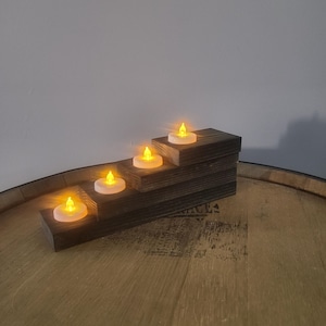 May include: A wooden candle holder with four LED candles. The holder is a dark brown, stepped design, and the candles emit a warm, yellow glow. The candles are placed on a wooden barrel.