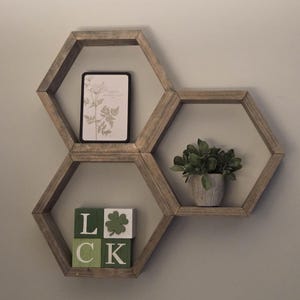 May include: Three wooden hexagon shelves mounted on a wall. The shelves hold a framed botanical print, a small potted plant, and wooden blocks spelling "LUCK" with a shamrock symbol. The shelves are made of light-colored wood.