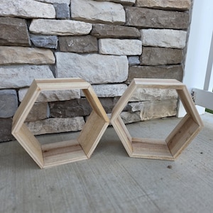 May include: Two unfinished wooden hexagon shelves. The shelves are made of light-colored wood and are connected. The shelves are sitting on a concrete surface in front of a stone wall.