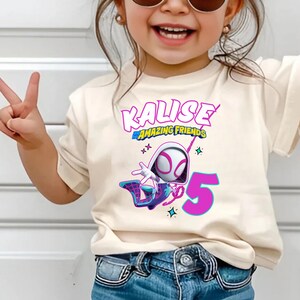 May include: A cream-colored t-shirt featuring a graphic of a superhero character, the name "Kalise" in pink, and the number "5" in magenta. The words "Amazing Friends" are also present.