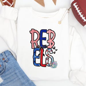 May include: White long-sleeved sweatshirt with the word "REBELS" in a patriotic design. The letters are in red, white, and blue patterns, including a gingham print and stars. A small bow and hearts accent the design. A pair of denim shorts is visible.