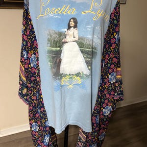 Puede incluir: Camiseta azul claro con un gráfico de Loretta Lynn con un vestido blanco y el texto "Loretta Lynn" en escritura amarilla. Las mangas son de tela azul marino con un estampado floral en rosa, azul y amarillo.