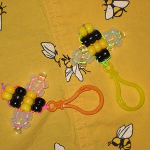 May include: Two beaded bee keychains. The bees are made with yellow and black beads, with clear wings. One keychain has an orange clasp, and the other has a yellow clasp. The background is yellow with bee illustrations.