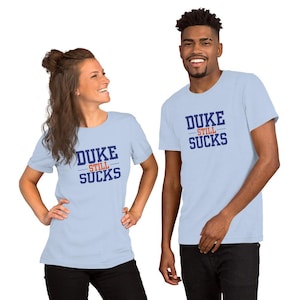 May include: Two light blue t-shirts with the text "DUKE STILL SUCKS" in navy blue and orange. The shirts are short-sleeved and worn by two people. The people are smiling and appear to be in a casual setting.