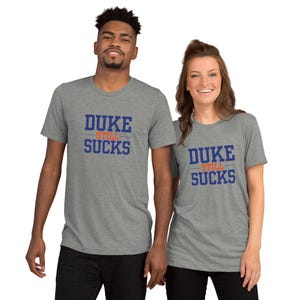 May include: Two people wearing gray t-shirts with the text "DUKE STILL SUCKS" in blue lettering. The shirts are short-sleeved and have a crew neck. The text is in a bold, collegiate-style font.