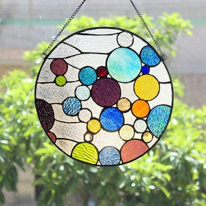 May include: A round stained glass window with a black metal frame and chain. The window features a design of colorful circles in shades of blue, yellow, orange, purple, red, and green. The glass has a textured appearance, and the background is blurred.