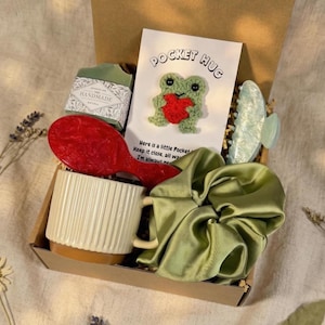 May include: A gift box containing a variety of items. Includes a red hairbrush, a white mug with a brown base, a green satin scrunchie, a green bar of soap, a light blue hair clip, and a card with a crocheted frog and the words "Pocket Hug".