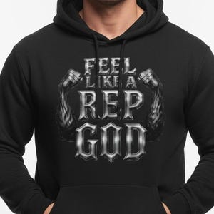 Könnte beinhalten: Schwarzer Kapuzenpullover mit dem Text "FEEL LIKE A REP GOD" in silberner, stilisierter Schrift. Das Design beinhaltet Bilder von Hanteln. Der Hoodie hat eine Vordertasche und eine Kapuze mit Kordelzug.