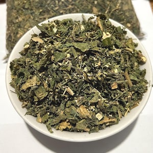 May include: A white bowl filled with a heap of dried, crushed green leaves and small woody stems. The mixture consists of various shades of dark and light green foliage, with some tan-coloured fragments visible throughout the textured herbal blend.