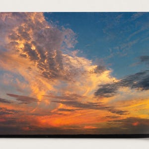 May include: A vibrant sunset sky featuring a dramatic display of golden, orange, and fiery red clouds contrasting against deep blue patches. The horizon line is dark and flat, emphasizing the expansive, textured cloud formations above.