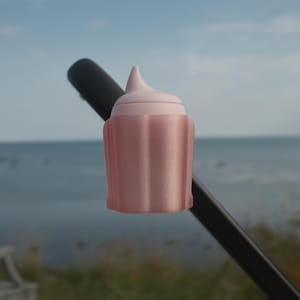 Könnte beinhalten: Ein hellrosa Flaschenverschluss mit dekorativem Design. Der Verschluss hat eine abgerundete Basis und eine konische Spitze. Der Verschluss wird von einer schwarzen Stange vor dem Hintergrund von Meer und Himmel gehalten.