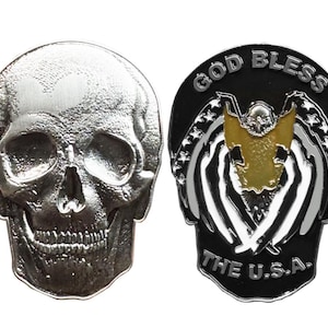 May include: A silver-colored skull coin with detailed facial features. The reverse side features a black background with the text "GOD BLESS THE U.S.A." and a gold-colored eagle design with white wings.