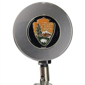 May include: A silver retractable badge holder with a circular design. The center features the National Park Service emblem, showcasing a mountain, tree, and bison. The text "NATIONAL PARK SERVICE" is displayed above the emblem.