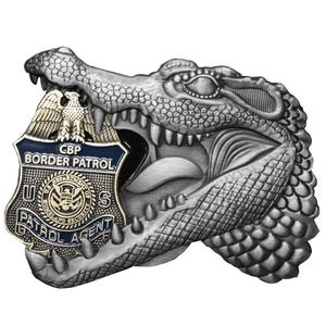 May include: A silver-colored CBP Border Patrol badge, featuring a detailed alligator design. The badge is held in the alligator's mouth. The badge has the text "CBP Border Patrol" and "Patrol Agent". The badge has a blue border and a gold eagle.