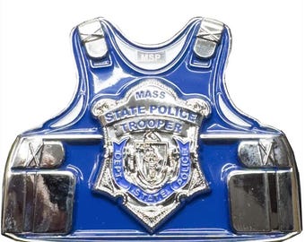 May include: A blue and silver metal badge in the shape of a police vest. The badge features the words "MASS STATE POLICE TROOPER" and a crest. The badge has a shiny, reflective surface.
