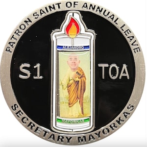 May include: A silver and black coin with the text "PATRON SAINT OF ANNUAL LEAVE SECRETARY MAYORKAS". The coin features an image of a person inside a lighter with the text "ALEJANDRO" and "MAYORKAS".