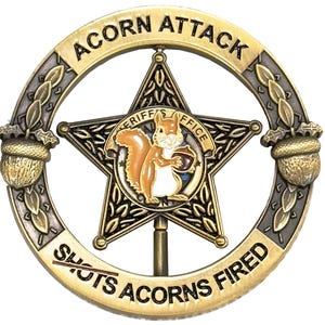 May include: A gold-toned metal badge with the words "ACORN ATTACK" and "SHOTS ACORNS FIRED" encircling a star-shaped emblem. The emblem features a cartoon squirrel holding an acorn, with "SHERIFF'S OFFICE" text.