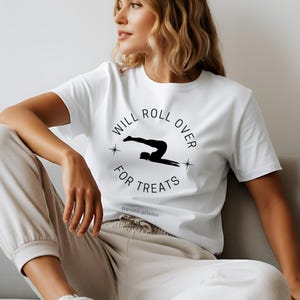 Puede incluir: Camiseta blanca con un gráfico negro de una persona haciendo ejercicio de pilates, rodeada por las palabras "WILL ROLL OVER FOR TREATS". El texto "@proto_pilates" está debajo. La persona lleva un pantalón deportivo beige.