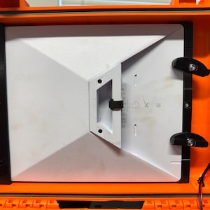 Puede incluir: Una maleta naranja impermeable abierta con un dispositivo electrónico blanco en su interior. El dispositivo se sujeta con soportes y cierres negros. Se ven cables rojos y negros.