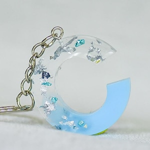 May include: A clear resin letter "C" keychain with silver and teal flakes. The bottom half of the letter is light blue. The keychain has a silver chain and key ring.