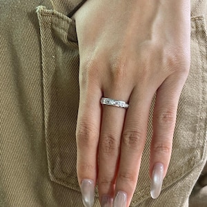 Può includere: Un anello d'argento con una fila di piccole pietre trasparenti è indossato su un dito. L'anello è posto su uno sfondo di tessuto beige. La mano della persona è visibile, con unghie lunghe e chiare.