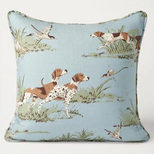 Könnte beinhalten: Quadratisches Zierkissen mit hellblauem Hintergrund, das eine Jagdszene zeigt. Das Design umfasst braune und weiße Hunde, Vögel und grünes Laub. Das Kissen hat einen hellblauen Rand.