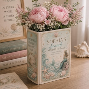 May include: A rectangular book-shaped vase featuring a light blue and cream color palette with illustrations of a mermaid, seashells, and starfish. The text reads Sophia's Seaside Mermaid Tale, Legends of the Tides and Timeless Dreams.