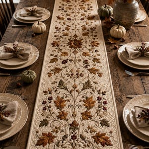 May include: A long, rectangular cream-colored table runner featuring an intricate quilted pattern of autumn leaves, acorns, and swirling vines in shades of brown, orange, gold, and green, centered on a rustic wooden dining table with place settings.