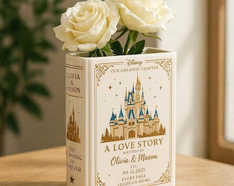 May include: A white ceramic book-shaped vase featuring a gold Disney castle illustration, the text "A LOVE STORY WRITTEN BY OLIVIA & MASON EST. 04.12.2025", and two white roses. The spine reads "OLIVIA & MASON" and "THE BEGINNING OF FOREVER".