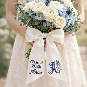 May include: A floral bouquet featuring white roses, blue hydrangeas, and foliage, secured with a large ivory bow. The bow displays the text "Class of 2026 Anna" and a monogram "A" with a graduation cap. The bouquet is held by someone wearing a white dress.
