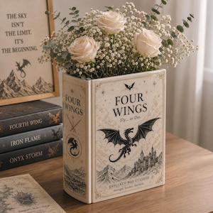 May include: A cream-colored ceramic book-shaped vase featuring a black illustration of a dragon, a castle, and the text "FOUR WINGS Fly... or Die. BASGIATH WAR COLLEGE Strength. Loyalty. Survival." filled with white roses and eucalyptus.