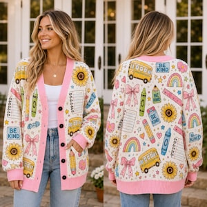 May include: A cream-colored knit cardigan featuring a repeating pattern of yellow school buses, sunflowers, pink bows, rainbows, crayons, and notebooks. The garment has a pink button-down placket, pink cuffs, and text that reads BE KIND.