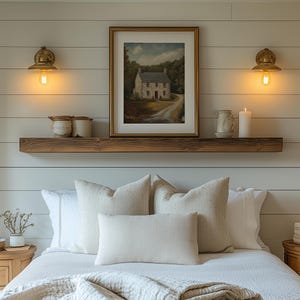 Puede incluir: Decoración de dormitorio con una pintura de paisaje enmarcada sobre una cama con almohadas y ropa de cama blancas. Una estantería rústica de madera contiene tarros decorativos, una taza y una vela. Dos apliques de pared de latón con bombillas Edison flanquean la pintura.
