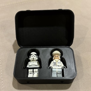 May include: A black plastic storage case with a hinged lid containing two small plastic figures. One figure is a white stormtrooper with black details, and the other is a figure wearing a white uniform with a hat and a brown moustache.