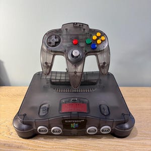 May include: A transparent gray Nintendo 64 console and controller. The console has the text "NINTENDO 64" on the front. The controller has colored buttons and a joystick. The console and controller are on a wooden surface.