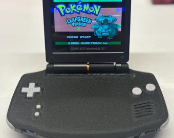 May include: A black Game Boy Advance SP handheld gaming console with a textured finish. The screen is powered on, displaying the title screen for Pokémon LeafGreen Version, featuring the text Press Start and the 2004 Game Freak Inc. copyright.