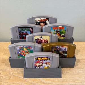 May include: A collection of Nintendo 64 video game cartridges, including titles like "Super Mario 64", "The Legend of Zelda", and "Star Fox 64". The cartridges are displayed in gray rectangular holders, showcasing the game labels.