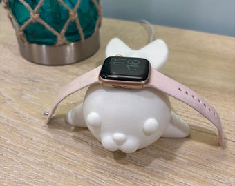 May include: A rose gold smartwatch with a light pink band rests on a white seal-shaped charging stand. The watch displays the time as 11:16. A turquoise glass lamp with a rope net is in the background.